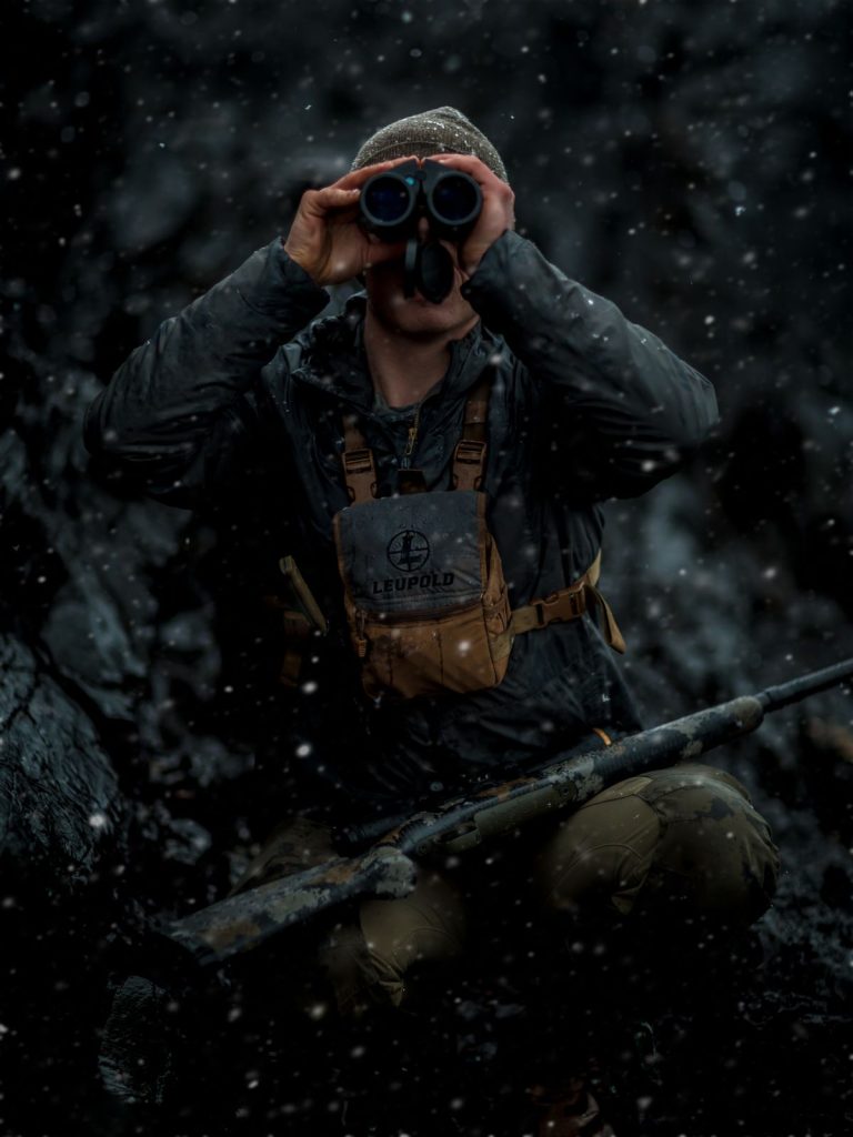 Hunter in falling snow glassing through binoculars, wearing a chest harness with a rifle across the knees