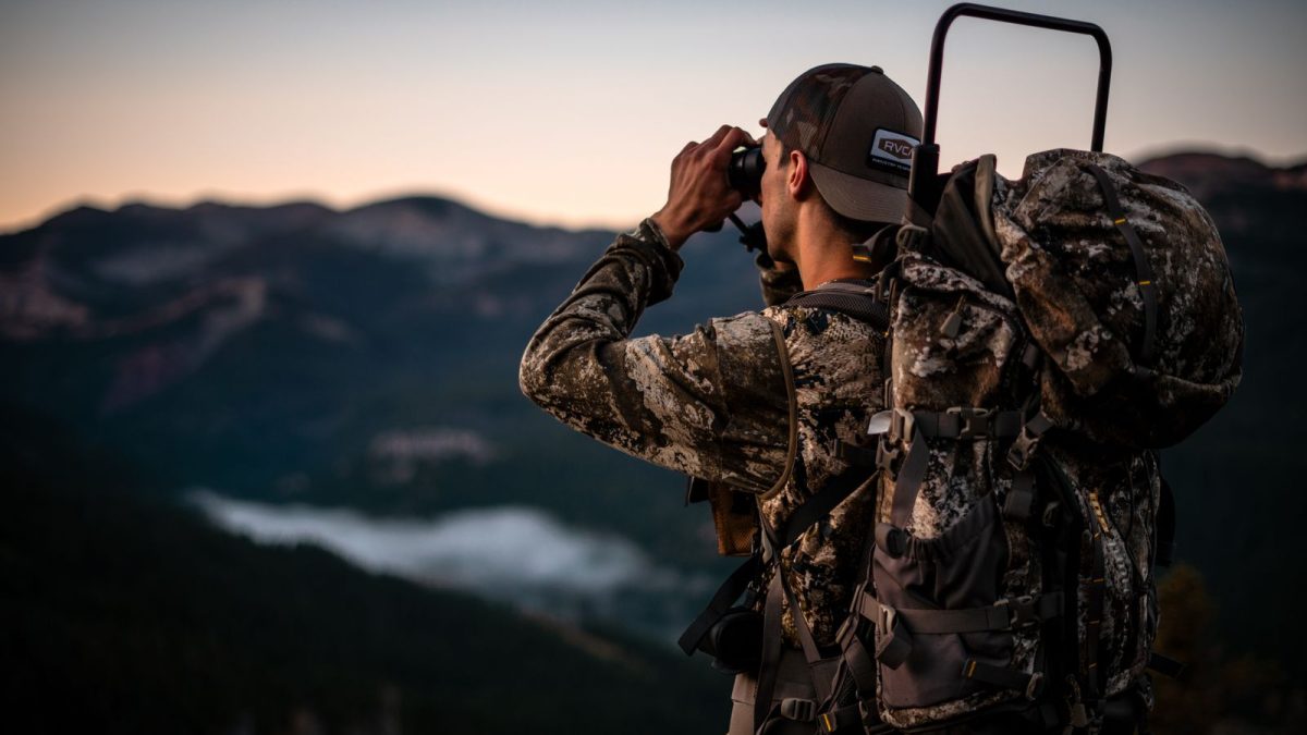 Camouflaged hunter with a pack glassing a mountain valley through binoculars at dusk
