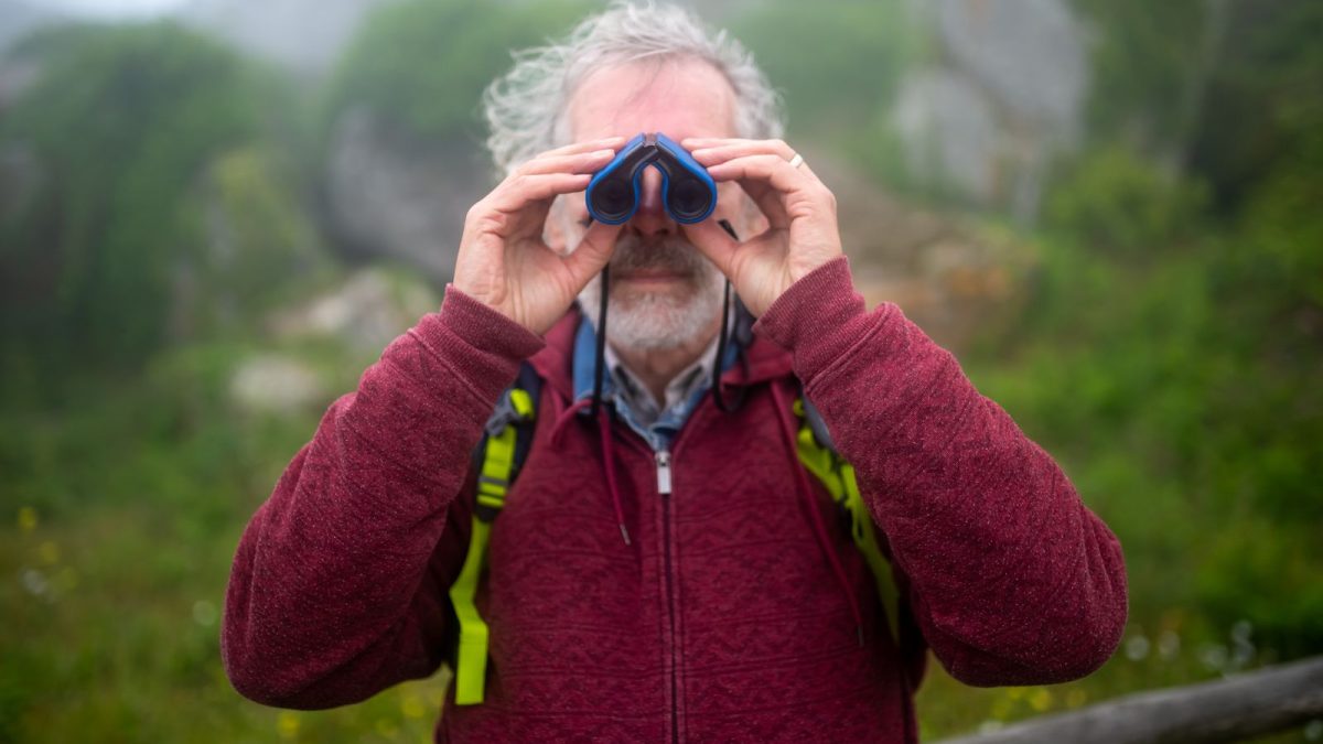 Hiker in a red sweater looking through binoculars on a misty green hillside